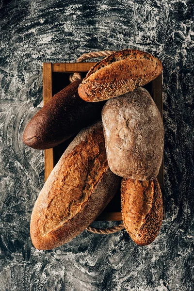 Blick von oben auf arrangierte Brotlaibe in Holzkiste auf dunkler Tischplatte mit Mehl — Stockfoto