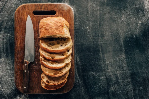 Draufsicht auf arrangierte Ciabatta-Stücke auf Schneidebrett mit Messer auf dunkler Oberfläche mit Mehl — Stockfoto