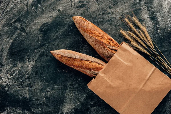 Vista superior de las baguettes francesas dispuestas en bolsa de papel y trigo sobre mesa oscura con harina - foto de stock