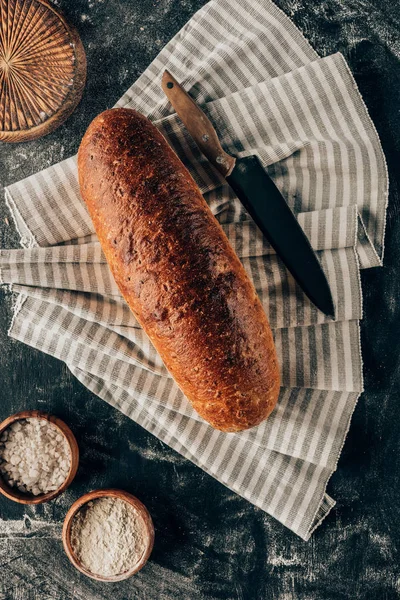 Piatto giaceva con pagnotta di pane su biancheria e ciotole con farina vicino sul tavolo scuro — Foto stock
