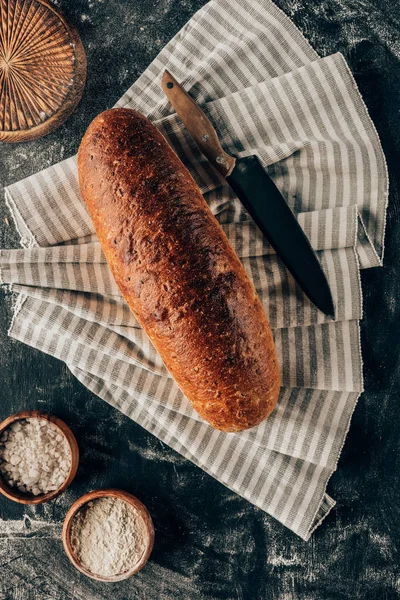 Flat lay with loaf of bread on linen and bowls with flour near by on dark tabletop — Stock Photo