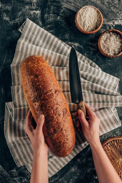 Partial view of female hands, knife and loaf of bread on linen on dark surface with flour — Stock Photo