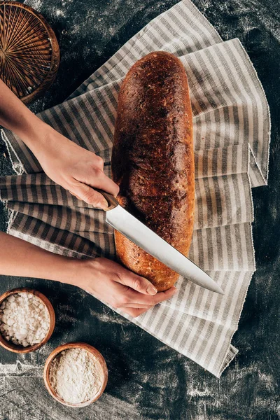 Cropped shot of woman cutting loaf of bread on linen on dark tabletop with flour — Stock Photo