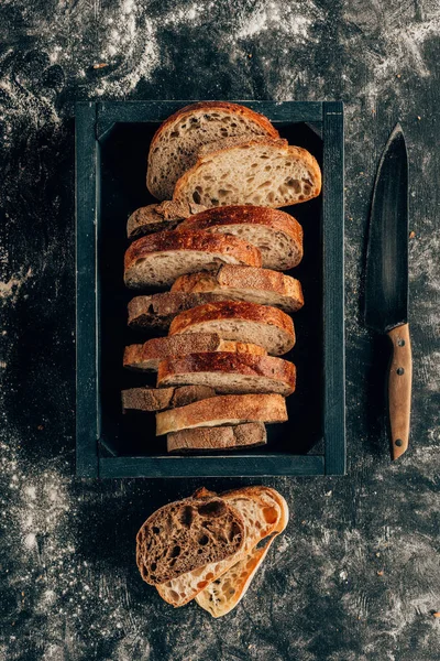 Top view of arranged pieces of bread in wooden box and knife on dark tabletop with flour — Stock Photo