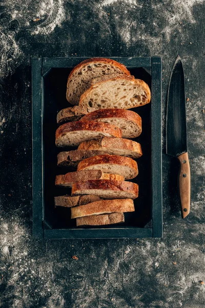 Top view of arranged pieces of bread in wooden box and knife on dark tabletop with flour — Stock Photo