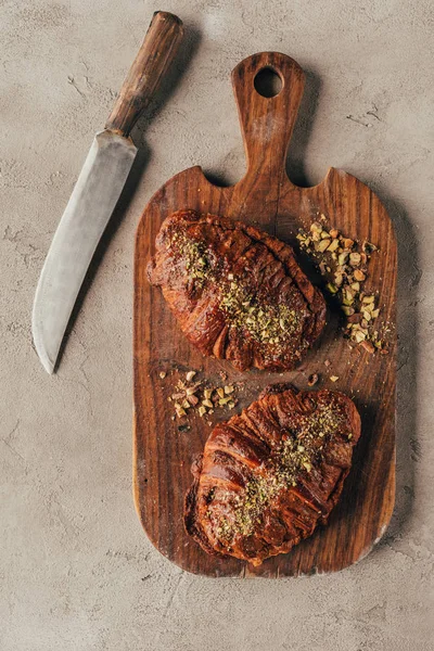 Top view of freshly baked croissants with pistachio nuts on wooden cutting board and knife on light surface — Stock Photo