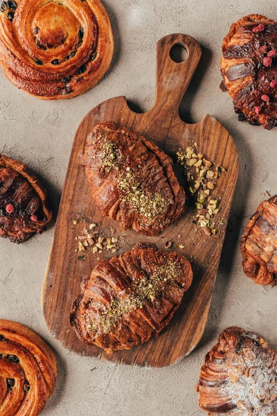 Flat lay with freshly baked sweet croissants with pistachio nuts on wooden cutting board on light surface — Stock Photo