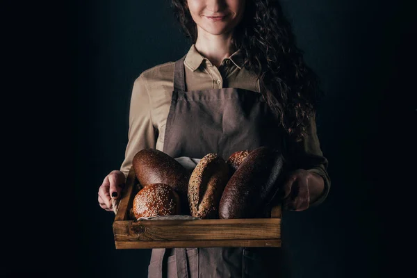 Cropped shot of smiling woman holding wooden box with loafs of bread in hands isolated on black — Stock Photo