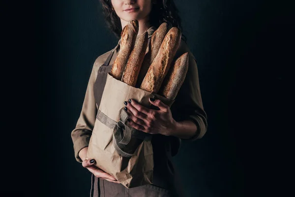 Tiro recortado de mujer en delantal sosteniendo baguettes francesas en bolsa de papel en manos aisladas en negro - foto de stock