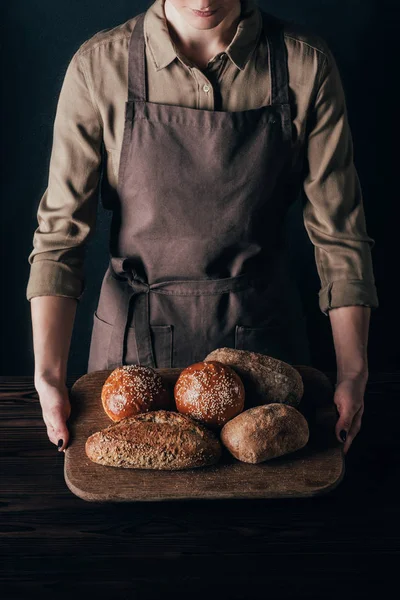 Partial view of woman holding loafs of bread on wooden cutting board isolated on black — Stock Photo