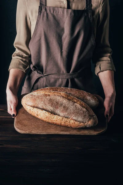 Partial view of woman holding loafs of bread on wooden cutting board isolated on black — Stock Photo