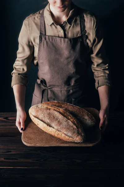 Partial view of woman holding loafs of bread on wooden cutting board isolated on black — Stock Photo