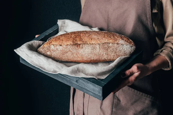 Cropped shot of woman holding wooden box with loaf of bread isolated on black — Stock Photo