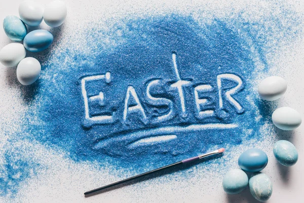 Top view of easter sign made of blue sand, brush with painted eggs on white — Stock Photo