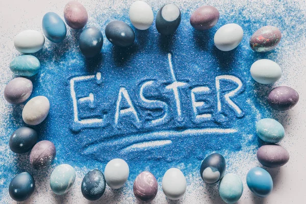 Вид зверху на Великодній знак з блакитного піску з пофарбованими яйцями на білому — стокове фото