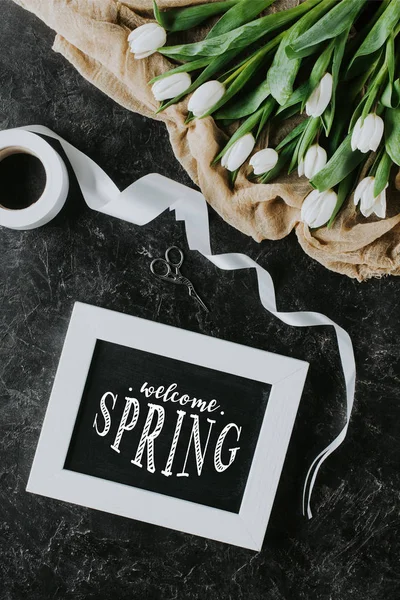 Vue de dessus des tulipes blanches, ruban et inscription BIENVENUE PRINTEMPS dans le cadre sur la surface noire — Photo de stock