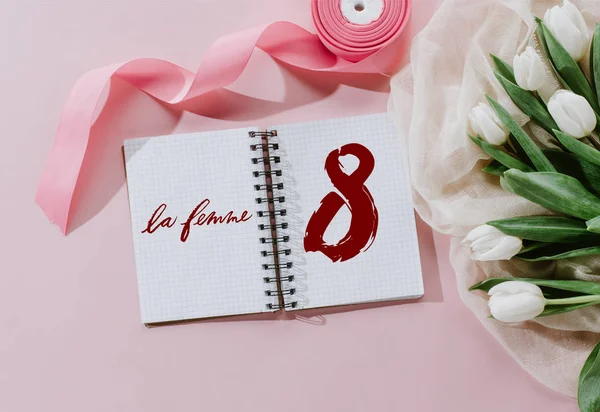 Ver en la parte superior del cartel de LE FEMME en bloc de notas, cinta rosa y tulipanes blancos para el día internacional de la mujer - foto de stock
