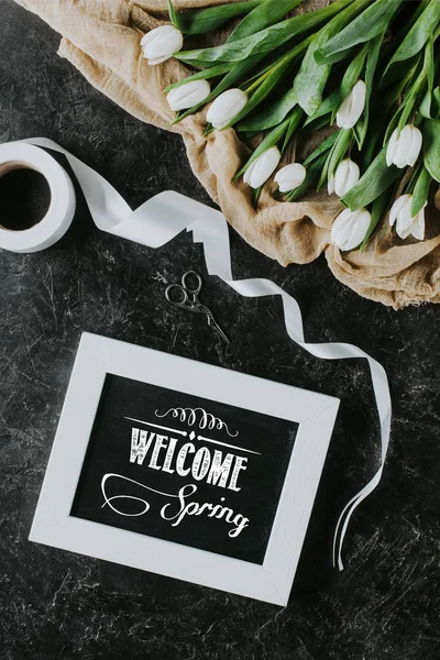 Вид зверху на білі тюльпани, стрічки та напис WELCOME SPRING в рамці на чорній поверхні — стокове фото