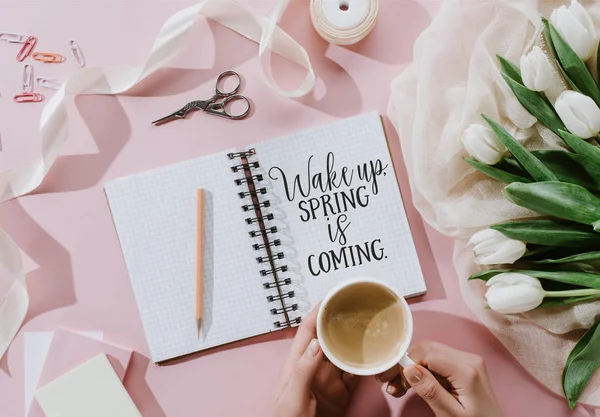 Vista recortada de la mujer sosteniendo taza de café en la superficie de color rosa con tulipanes y despertar. PRIMAVERA VIENE la inscripción en el bloc de notas - foto de stock