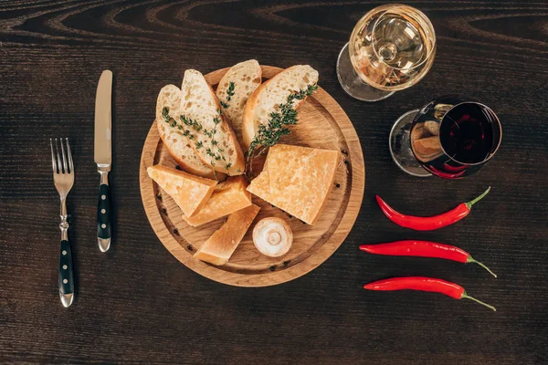 Top view of parmesan cheese with baguette slices on wooden board, chili peppers and wine on table — Stock Photo