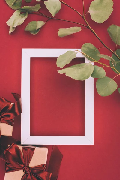 Вид зверху порожня біла рамка, подарункові коробки і гілочка з зеленим листям на червоному — стокове фото