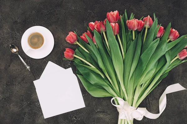 Vista dall'alto di bellissimi tulipani rossi, tazza di caffè e busta bianca con biglietto di auguri — Foto stock