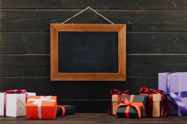 Leere Kreidetafel im Rahmen mit Geschenkboxen, die an der Holzwand hängen — Stockfoto