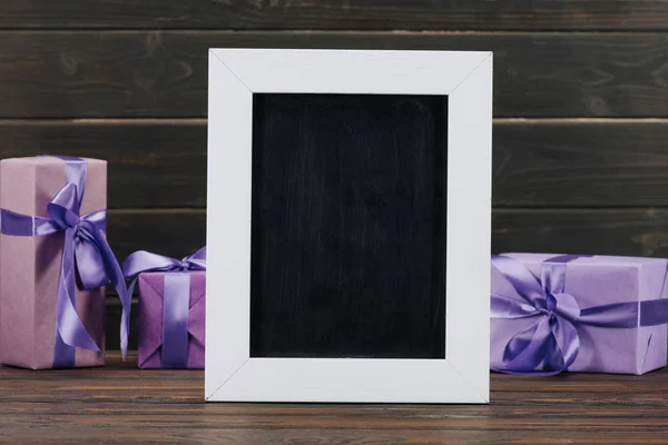 Blank chalkboard in frame with gift boxes against wooden wall — Stock Photo
