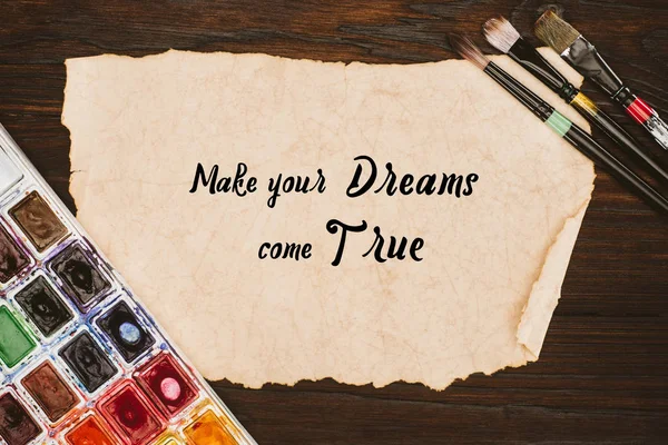 Top view of aged paper with make your dreams come true lettering and painting supplies on wooden table — Stock Photo