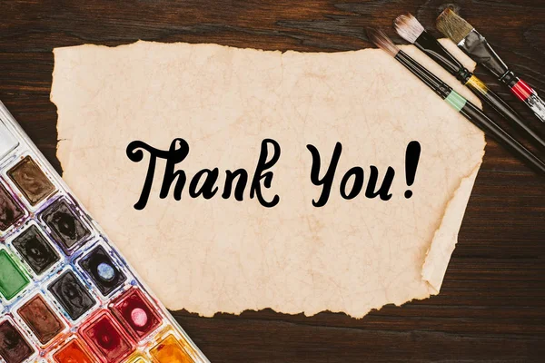 Top view of aged paper with thank you lettering and painting supplies on wooden table — Stock Photo