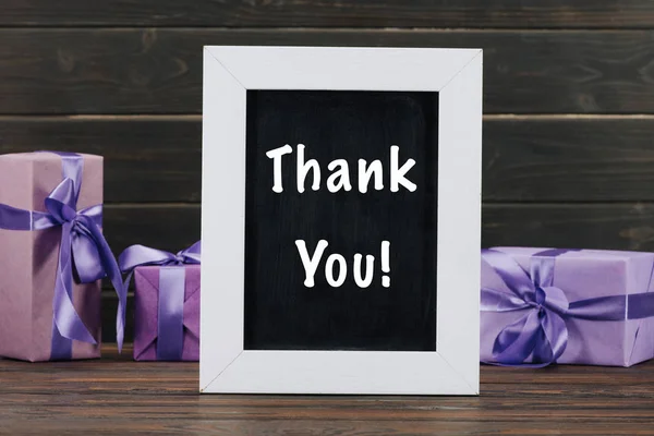 Thank you lettering on chalkboard in frame with gift boxes against wooden wall — Stock Photo