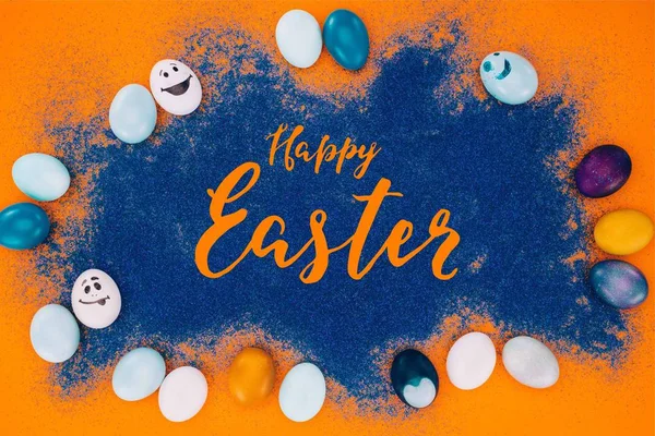 Top view of happy easter lettering on blue sand and easter eggs on orange surface — Stock Photo