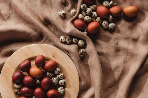 Vue du dessus des oeufs peints de Pâques et des oeufs de caille sur une assiette en bois et une nappe en lin — Photo de stock