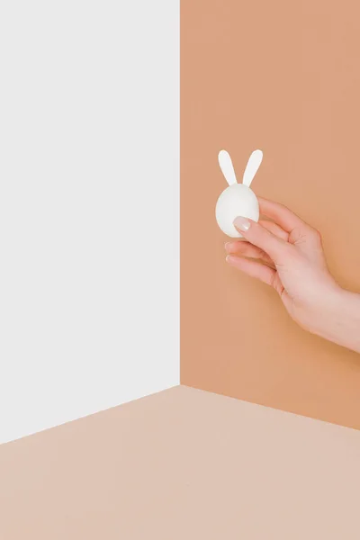 Обрізаний вид людини, що тримає пасхальне яйце з вухами кролика на бежевому та білому мінімалістичному фоні — стокове фото