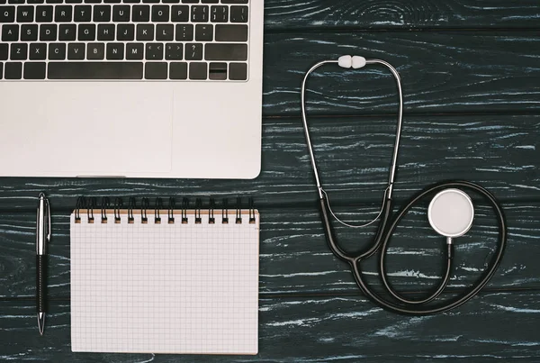 Top view of arranged laptop, empty notebook and stethoscope on dark wooden tabletop — Stock Photo