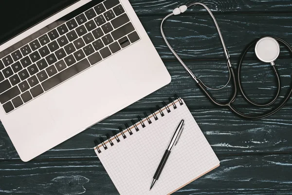 Top view of arranged laptop, empty notebook, pen and stethoscope on dark wooden tabletop — Stock Photo