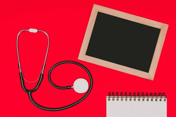 Top view of blank chalkboard, notebook and stethoscope isolated on red tabletop, world health day concept — Stock Photo