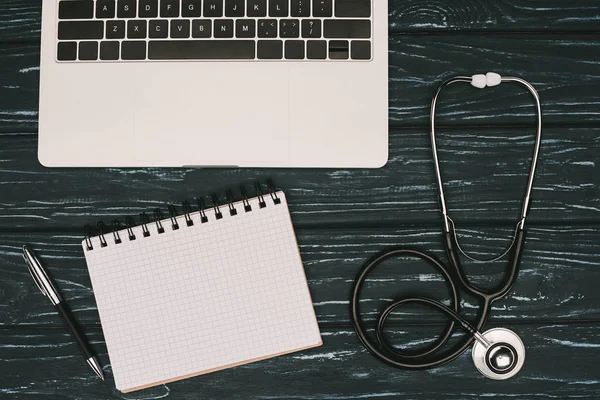 Top view of arranged laptop, empty notebook, pen and stethoscope on dark wooden tabletop — Stock Photo