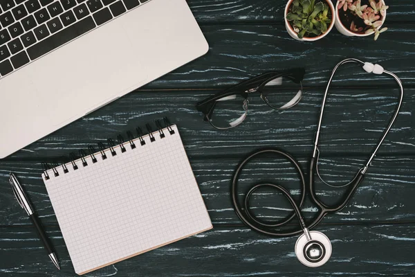 Top view of arranged laptop, empty notebook, eyeglasses and stethoscope on dark wooden tabletop — Stock Photo