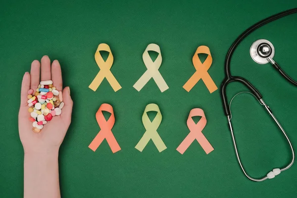 Tiro recortado de la mano femenina con píldoras, cintas coloridas dispuestas y estetoscopio aislado en la mesa verde, concepto del día de la salud mundial - foto de stock