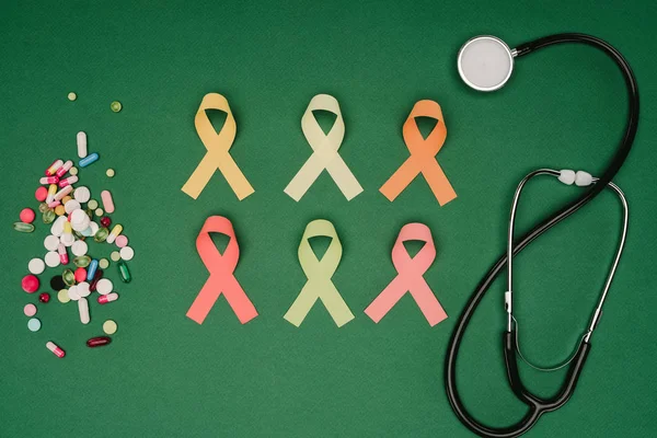 Acostado plano con píldoras arregladas, cintas de colores y estetoscopio aislado en la mesa verde, concepto del día de la salud mundial - foto de stock