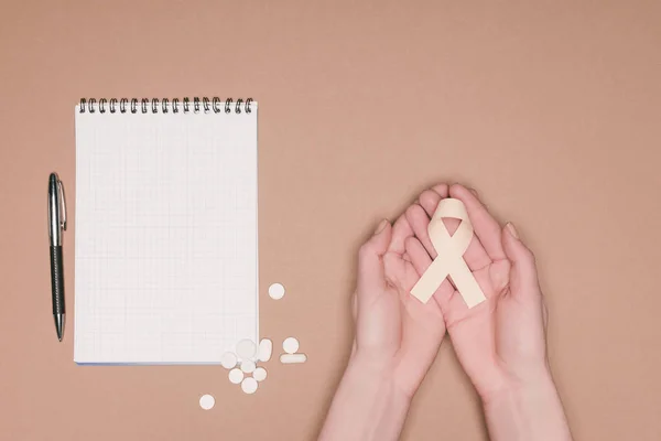 Cropped shot of female hands with ribbon, pills and empty notebook isolated on beige tabletop, world health day concept — Stock Photo