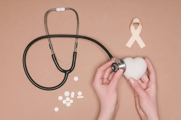 Vista parziale delle mani femminili con cuore, nastro rosa e stetoscopio isolati sulla superficie beige, concetto giornata mondiale della salute — Foto stock