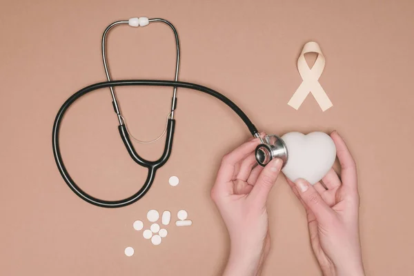 Vue partielle des mains féminines avec cœur, ruban rose et stéthoscope isolés à la surface beige, concept de journée mondiale de la santé — Photo de stock