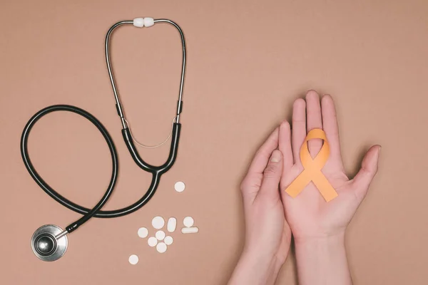 Обрезанный снимок женских рук с лейкемией, раком почек, рассеянным склерозом лента осознания RSD, таблетки и стетоскоп, изолированные на бежевом столе — стоковое фото