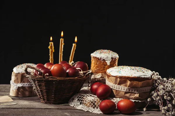 Bougies allumées, œufs peints dans le panier et gâteaux de Pâques savoureux sur noir — Photo de stock