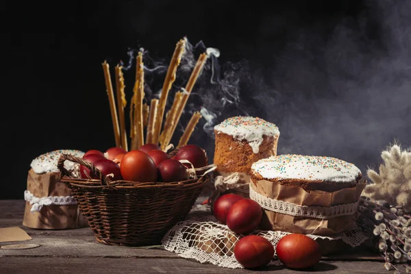 Раскрашенные яйца в корзину, свечи с дымом и домашние пасхальные торты на деревянном столе — стоковое фото
