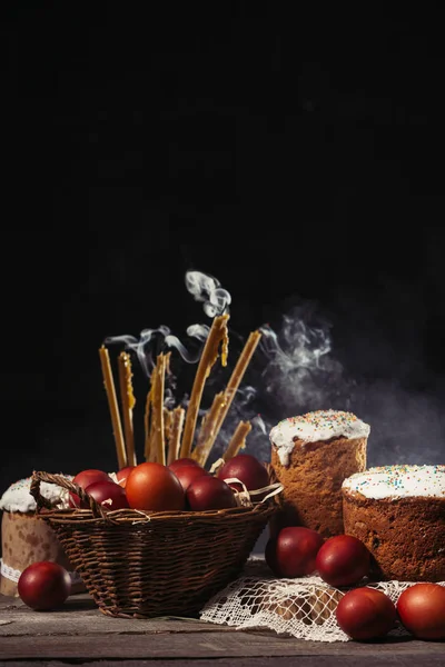 Candles with smoke, homemade easter cakes and painted chicken eggs in basket on wooden table — Stock Photo