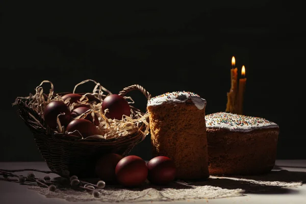 Basket with painted chicken eggs, easter cakes and burning candles on black — Stock Photo