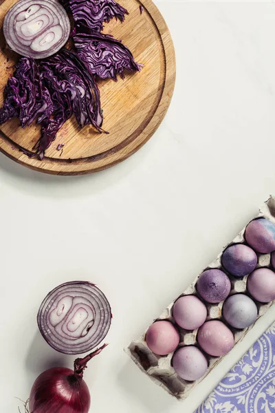 Верхний вид окрашенных пасхальных яиц в яичную коробку, фиолетовая капуста и лук на сером — стоковое фото